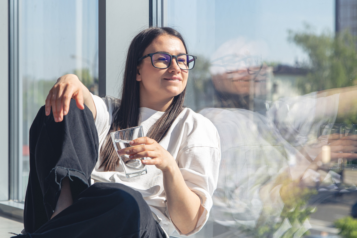 Une jeune femme joyeuse boit de l'eau assise près de la fenêtre un matin d'été