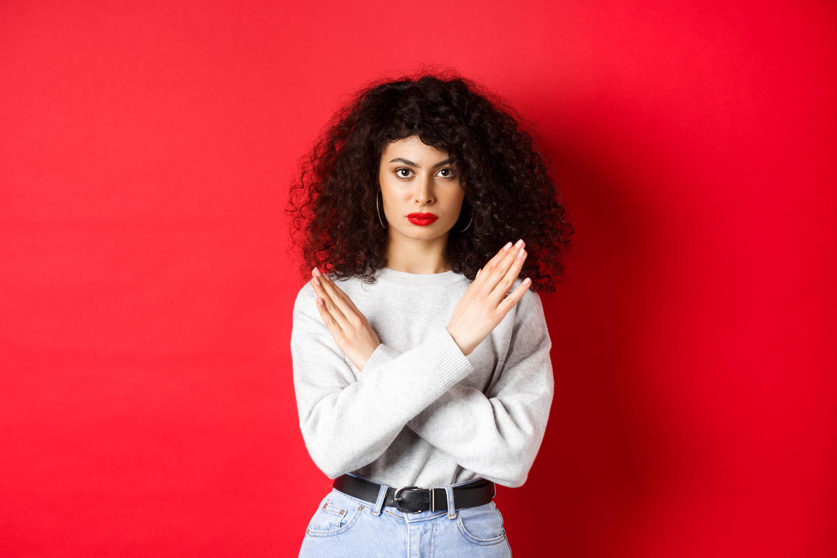 Une étudiante femme caucasienne sérieuse aux cheveux bouclés dit non, faisant un geste croisé pour arrêter ou interdire quelque chose de mauvais, se tenant mécontente sur fond rouge.