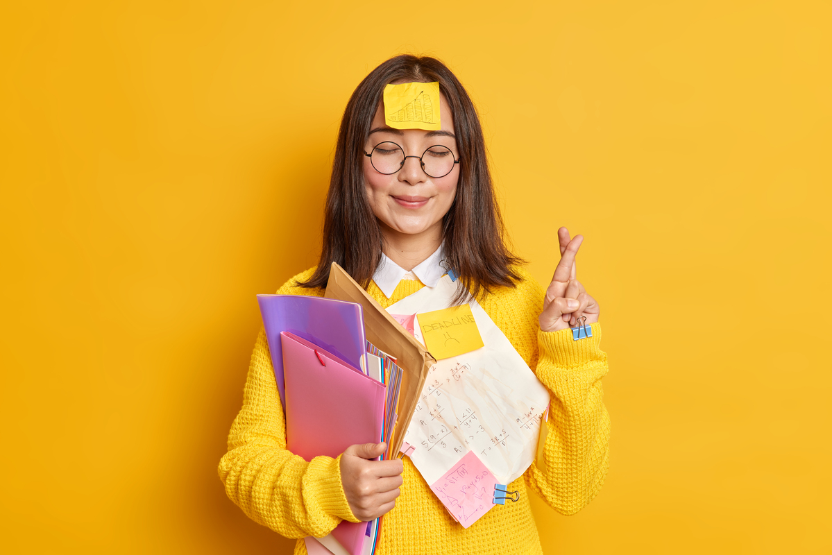 étudiante qui souris avec pleins de post-it, qui tient des classeurs et qui croise les doigts