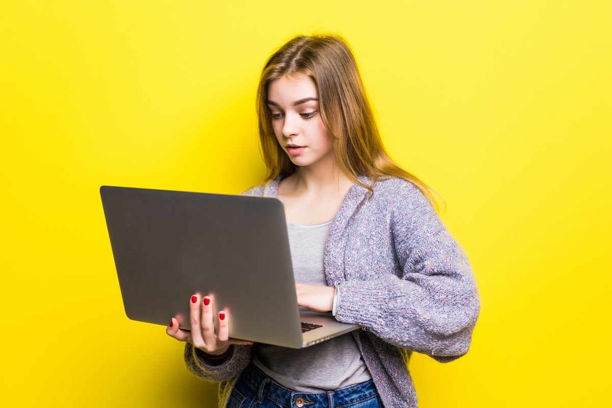 Portrait of a smiling teenage girl holding laptop computer isolated on a yellow background