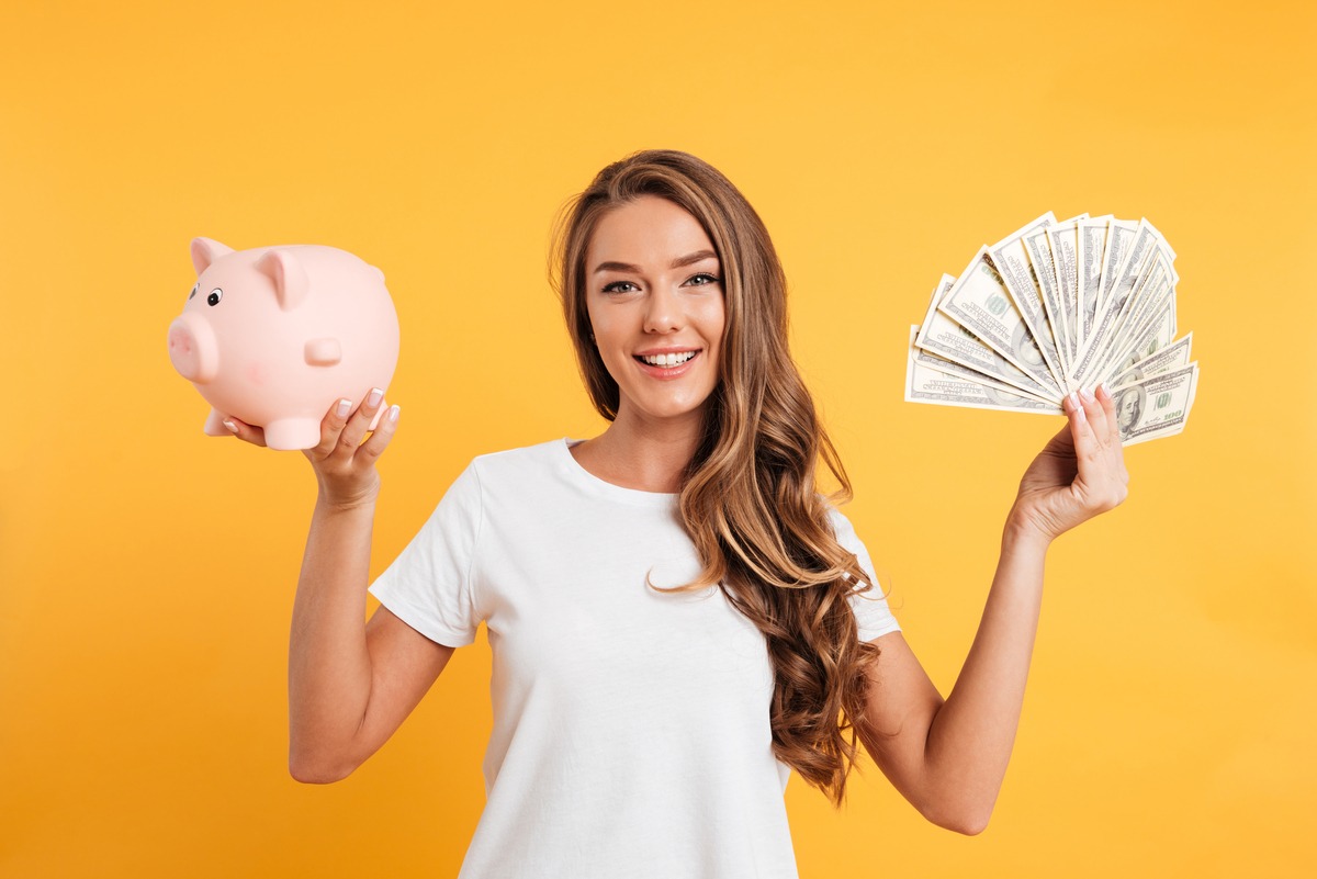 Portrait of a cheery young girl holding piggy bank and bunch of money banknotes isolated over yellow background