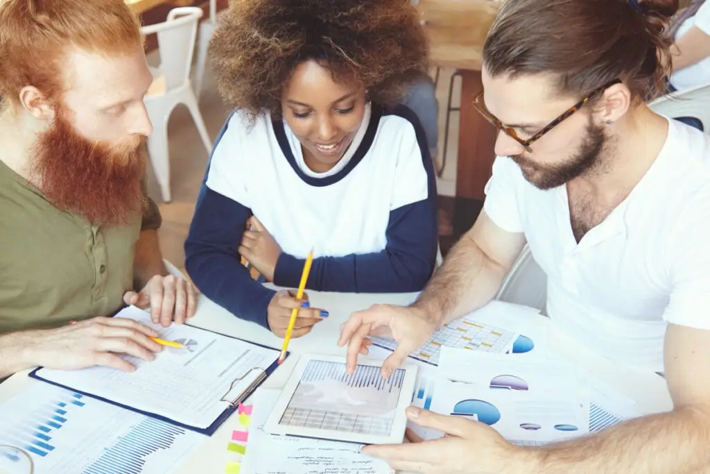 Groupe de trois jeunes spécialistes du marketing du futur, intelligents et intelligents, travaillant ensemble sur un projet de fin d'études, analysant des graphiques et des tableaux, utilisant une tablette tactile.